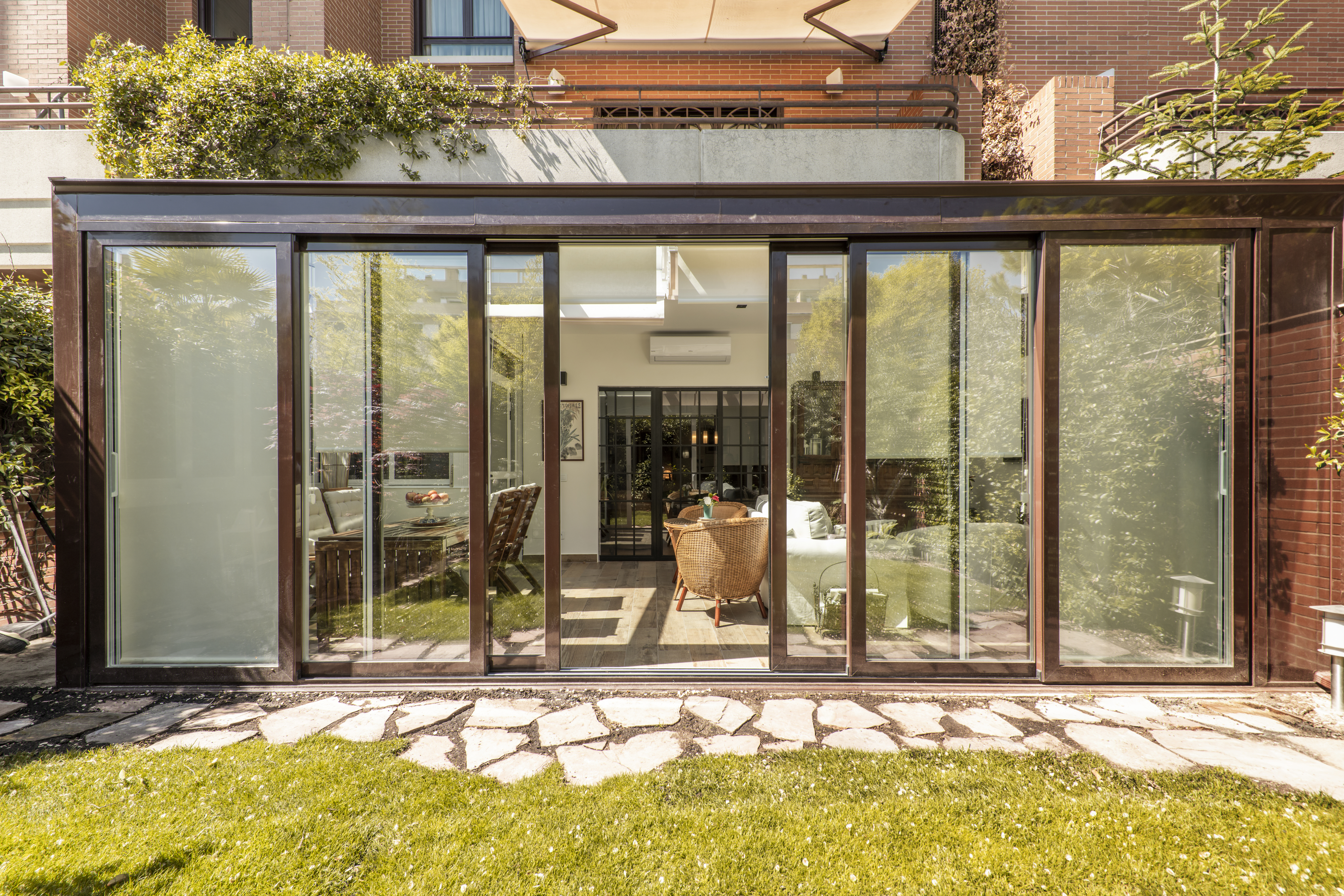 Covered terrace with brown aluminum structure and glass with access to a house on the ground floor and a garden with a path of stone slabs and grass